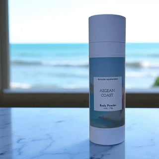 Body powder container labeled 'Aegean Coast' on a marble surface with a beach view in the background