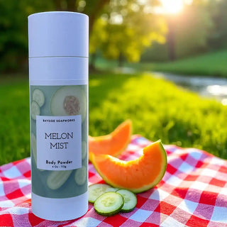 Body powder can labeled 'Melon Mist' on a checkered tablecloth with melon and cucumbers outdoors.