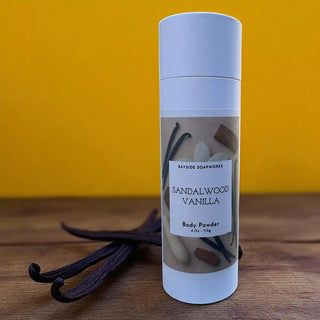 Body powder container labeled 'Sandalwood Vanilla' with vanilla beans on a wooden surface and yellow background