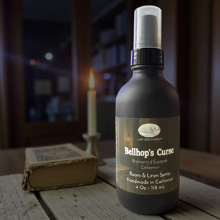 Room & Linen Spray bottle labeled 'Bellhop's Curse' by Lazy Dog Candle Co. on a wooden surface with a candle and book in the background.