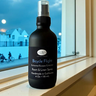 Black room and linen spray bottle labeled 'Bicycle Flight' on a windowsill with a cityscape in the background.