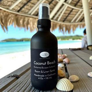 Black room and linen spray bottle labeled 'Coconut Beach' on a wooden surface with beach scenery in the background.