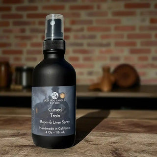 Black room and linen spray bottle labeled 'Cursed Train' by Lazy Dog Candle on a wooden surface with a brick wall background.