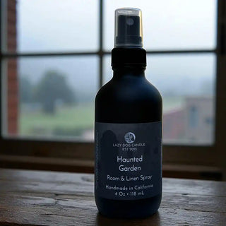 Room spray bottle labeled 'Haunted Garden' by Lazy Dog Candle on a wooden surface with a window in the background.