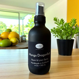 Black room & linen spray bottle labeled 'Mango Dragonfruit' on a wooden surface with a plant and fruit in the background.