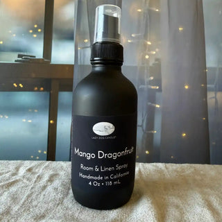 Black room & linen spray bottle labeled 'Mango Dragonfruit' on a textured surface with a blurred background.