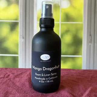 Black room & linen spray bottle labeled 'Mango Dragonfruit' on a red surface with a blurred green and white background.