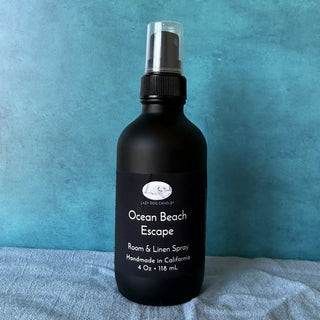 Black room & linen spray bottle labeled 'Ocean Beach Escape' on a textured blue surface.
