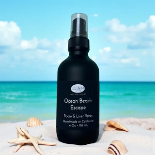 Black bottle of 'Ocean Beach Escape' room & linen spray on a beach with shells and ocean view.