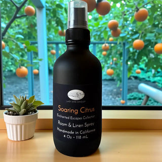 Black room and linen spray bottle labeled 'Soaring Citrus' on a wooden surface with an outdoor background.