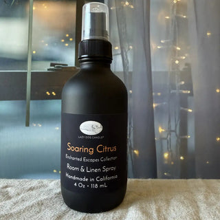 Black room spray bottle labeled 'Soaring Citrus' on a textured surface with blurred lights in the background.