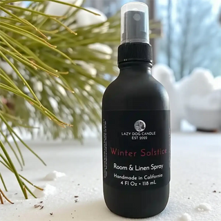 Black room & linen spray bottle labeled 'Winter Solstice' by Lazy Dog Candle on a snowy surface with plants.
